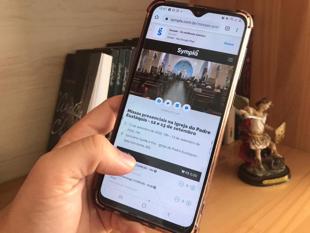 Mão com celular mostra a página do Sympla para retirada de ingressos para as missas presenciais.